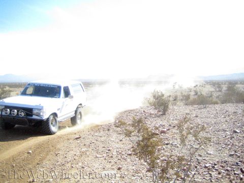 1990 Ford Bronco - white Ford Bronco with a light bar bumper drives fast along a dusty desert trail kicking up a dust cloud.