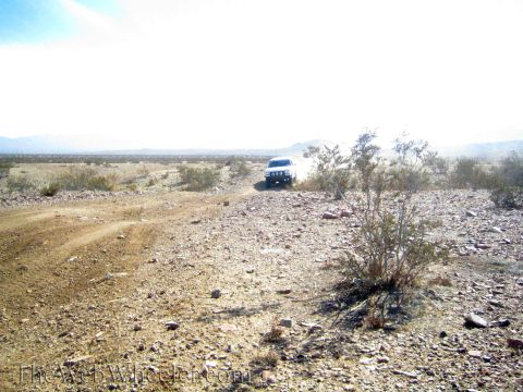 1990 Ford Bronco - distant white vehicle drives across an open rocky desert with low mountains on the horizon.