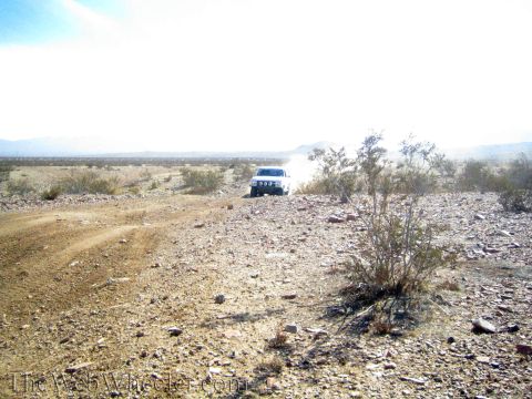1990 Ford Bronco - white vehicle moves across a rocky desert flat, raising dust under a bright hazy sky.