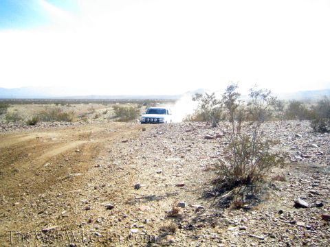 1990 Ford Bronco - white vehicle kicks up dust crossing rocky desert terrain with distant mountains beyond.