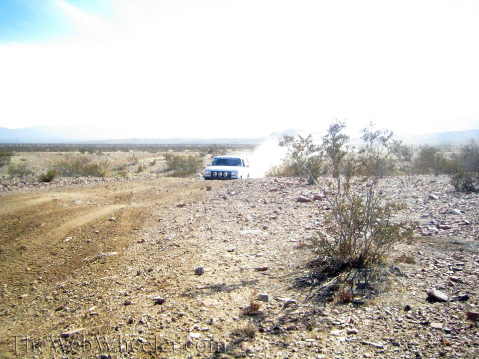 White vehicle kicking up dust on rocky desert terrain with mountains beyond