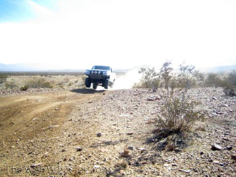 1990 Ford Bronco - dark lifted truck with a front light bar drives a dusty desert trail across rocky open terrain.