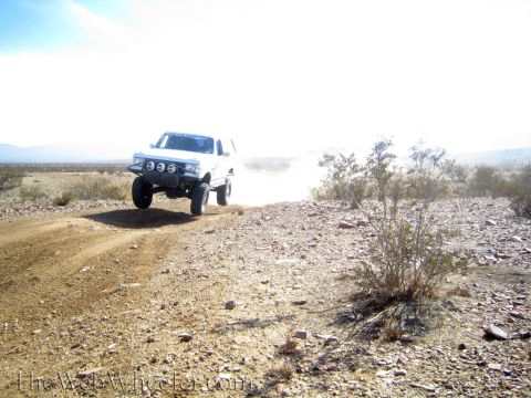 1990 Ford Bronco - white lifted truck with auxiliary lights drives toward the camera, raising dust on a desert trail.