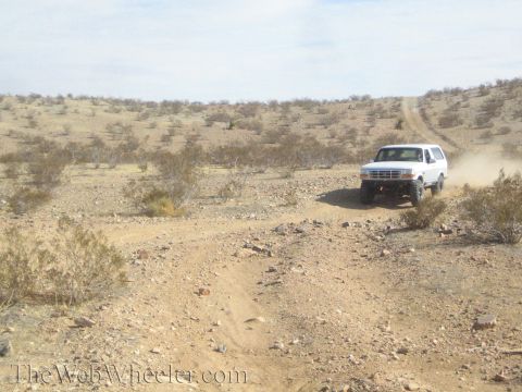 1990 Ford Bronco - white Ford Bronco drives along a desert two-track trail crossing brush-covered rocky scrubland.