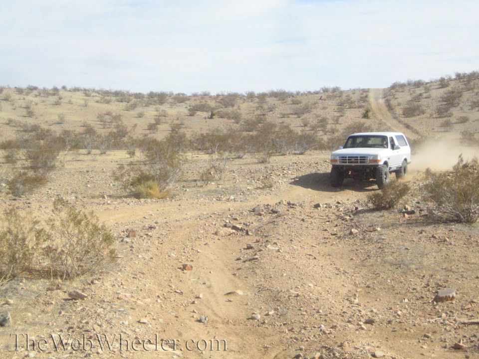 White Ford Bronco driving a desert two-track trail across scrubland