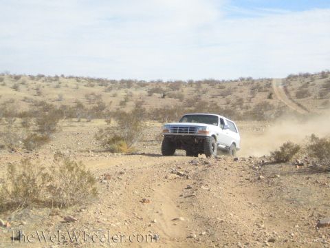 1990 Ford Bronco - white Ford Bronco rounds a curve on a dusty desert trail through open scrubland.