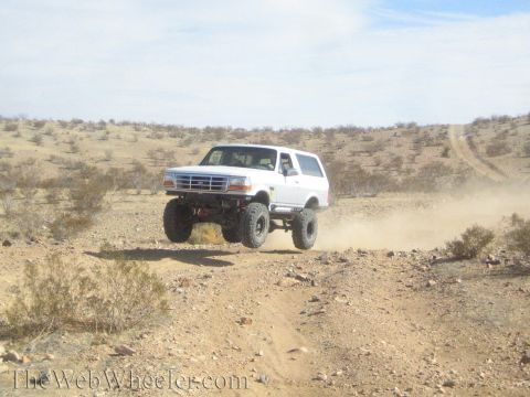1990 Ford Bronco - white lifted Ford Bronco catches air over a rise on a dusty desert trail, exposing its undercarriage.