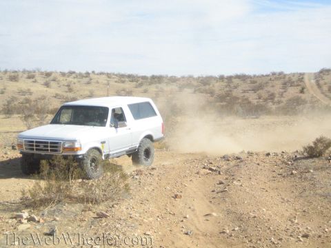 1990 Ford Bronco - white lifted Ford Bronco drives a dusty desert trail seen from the side, trailing dust across scrubland.