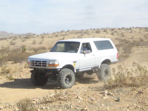 1990 Ford Bronco - white lifted Ford Bronco on large mud-terrain tires sits on a desert trail with a driver visible inside.