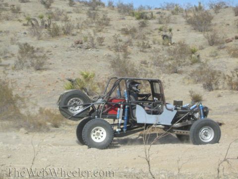 1990 Ford Bronco - bare tube-frame off-road buggy with a helmeted driver and rear-mounted spare tire drives a desert trail.