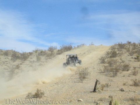 1990 Ford Bronco - off-road buggy climbs a steep dusty desert hill, kicking up a large dust cloud under a blue sky.