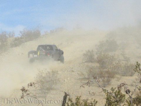 1990 Ford Bronco - dark off-road truck climbs a dusty desert hillside, partly enveloped in a thick cloud of dust.