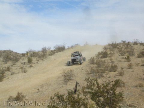 1990 Ford Bronco - off-road buggy descends a long dusty desert hillside trail amid scattered brush under a blue sky.