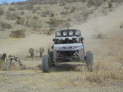 1990 Ford Bronco - silver tube-frame off-road buggy with a roof light bar climbs a desert hill past cacti and brush.