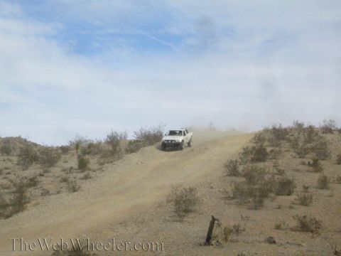 1990 Ford Bronco - white vehicle crests a dusty desert hill trail among low brush under a partly cloudy sky.