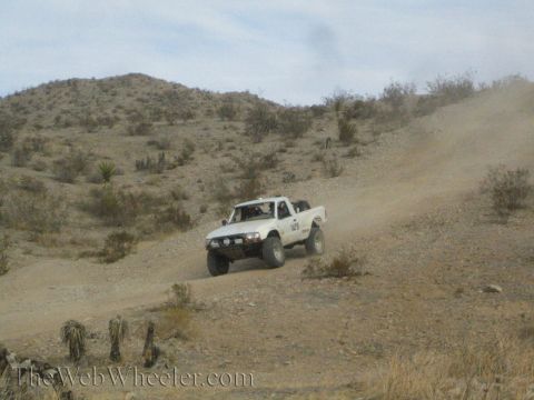 1990 Ford Bronco - white lifted off-road pickup climbs a steep dirt trail in a dry desert hill landscape.