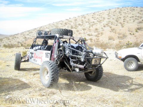 1990 Ford Bronco - single-seat tube-chassis off-road race buggy marked 167 sits in dry desert scrub beside a white SUV.