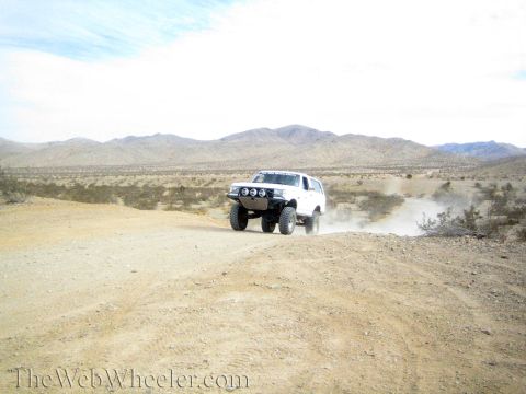1990 Ford Bronco - white lifted SUV with light bar speeds along a dirt road, raising dust against distant mountains.