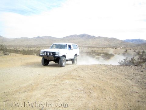 1990 Ford Bronco - white lifted full-size SUV with front light pods drives a dirt desert road, trailing dust.