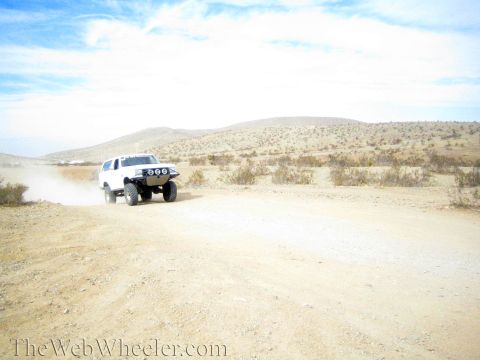 1990 Ford Bronco - white lifted SUV with front light pods kicks up dust on a broad dirt road under hazy sky.