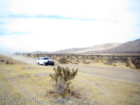 1990 Ford Bronco - white SUV travels a desert trail in the distance, raising dust amid open scrubland and mountains.
