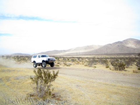 1990 Ford Bronco - white lifted SUV catches air over a dirt desert road, trailing dust against distant mountains.