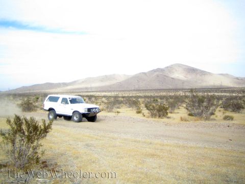 1990 Ford Bronco - white lifted full-size SUV rounds a bend on a dirt desert road below tan hills.