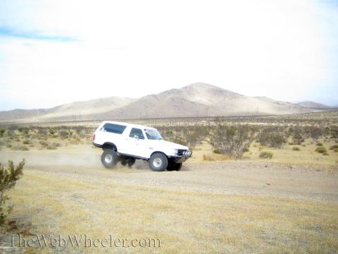 1990 Ford Bronco - white lifted SUV drives a desert trail, throwing up a dust cloud beneath a tan mountain.