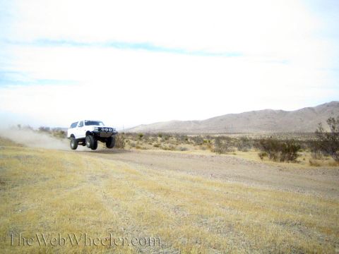 1990 Ford Bronco - white lifted SUV with front lights races along a dirt road, leaving a dust trail in the desert.