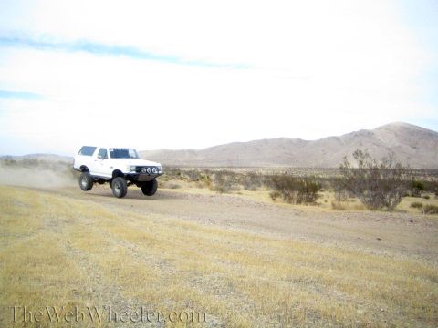 1990 Ford Bronco - white lifted SUV speeds down a dirt road in the desert, trailing dust below a tan ridge.