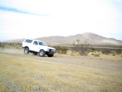 1990 Ford Bronco - white lifted SUV leans into a turn on a dirt desert road, kicking up dust near tan hills.