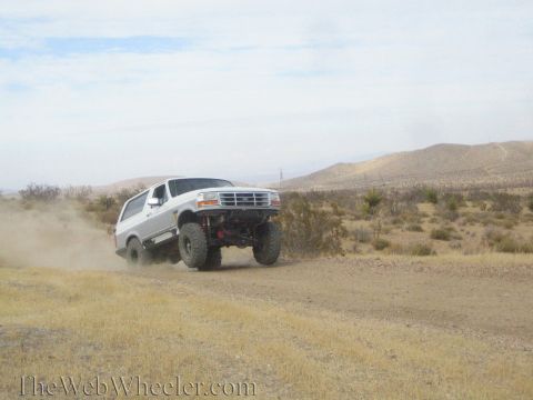 1990 Ford Bronco - white lifted Ford full-size SUV with Ford grille emblem jumps a dirt desert road, trailing dust.