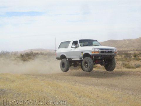 1990 Ford Bronco - white lifted Ford full-size SUV with Ford oval grille gets airborne over a dirt desert road.