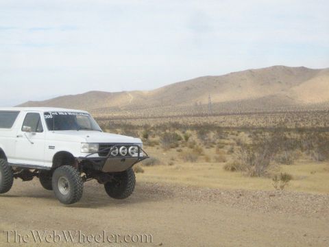 1990 Ford Bronco - white lifted full-size SUV with front light pods and bumper stickers sits on a desert ridge road.