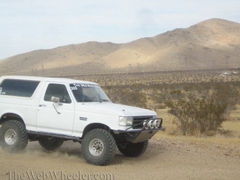 1990 Ford Bronco - white lifted full-size SUV with front light pods drives a dirt desert road beneath tan mountains.