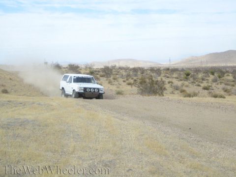 1990 Ford Bronco - white lifted SUV with front light pods rounds a curve on a desert trail, raising dust.