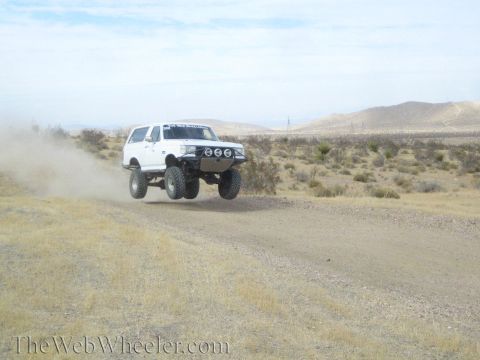 1990 Ford Bronco - white lifted SUV catches big air over a dirt road in the desert, trailing a dust cloud.