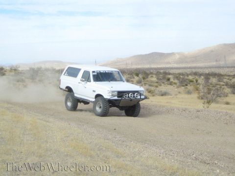 1990 Ford Bronco - white lifted full-size SUV with front light pods drives a dirt desert road below tan hills.