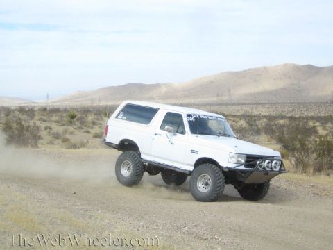 1990 Ford Bronco - white lifted full-size SUV with front light pods leans into a turn on a dirt desert road.