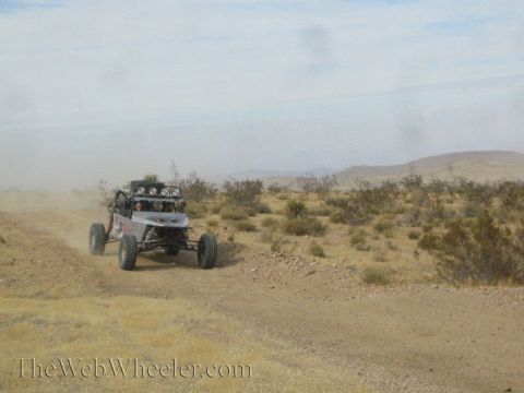 1990 Ford Bronco - tube-chassis off-road buggy with rear light bar speeds down a dusty desert trail away from camera.