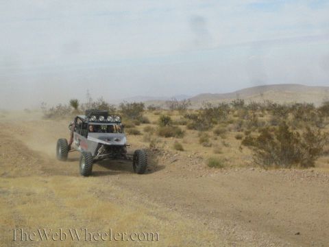 1990 Ford Bronco - tube-chassis off-road race buggy rounds a bend on a dusty desert trail amid scrub.