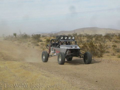1990 Ford Bronco - single-seat tube-chassis off-road race buggy with roof light bar drives a dusty desert trail.