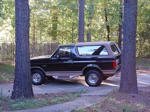 1994 Ford Bronco - A black Ford Bronco with a tan top and trim is parked on a concrete driveway, partially obscured by trees.