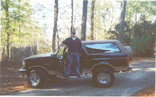 1994 Ford Bronco - A man poses with his Ford Bronco parked outdoors on a paved surface, surrounded by trees.