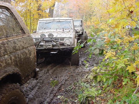 1994 Ford Bronco - Several off-road vehicles navigate a challenging muddy path surrounded by fall foliage.