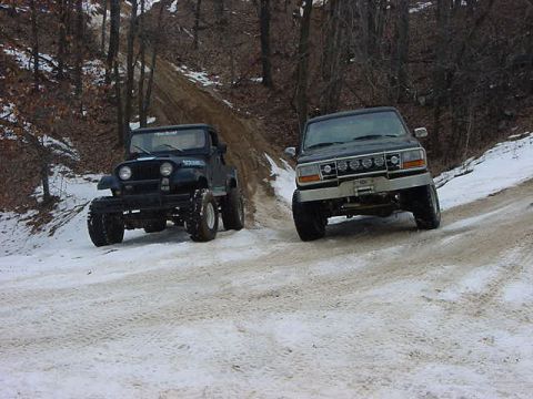 1994 Ford Bronco - Two off-road vehicles navigate a snowy path through a wooded area.