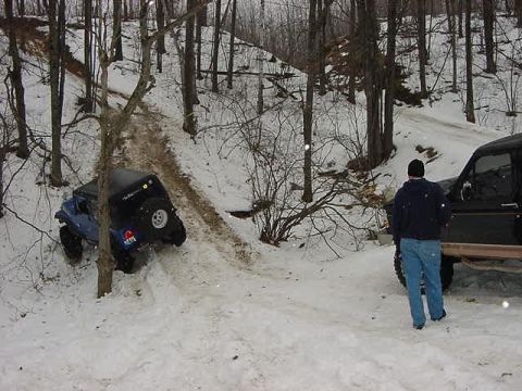 1994 Ford Bronco - Two off-road vehicles navigate a snowy, wooded trail, with one vehicle struggling to climb a steep incline.