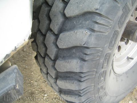 1990 Ford Bronco - Detailed close-up of a deeply lugged mud-terrain off-road tire mounted on a steel wheel, set against the white lower body of a truck.