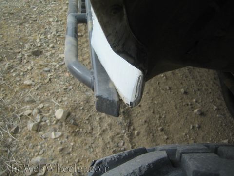 1990 Ford Bronco - cracked and split white plastic body panel hanging over a tubular rock-slider step, photographed against gravelly trail dirt.