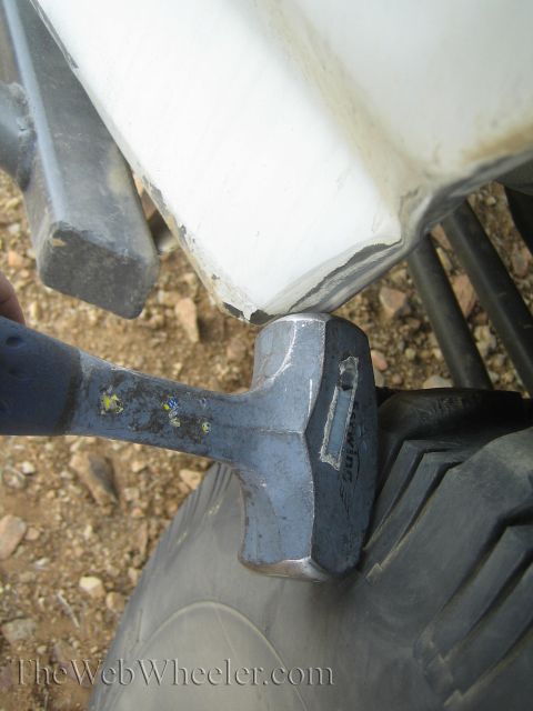 1990 Ford Bronco - heavy sledgehammer head propped against the cracked lower edge of a white truck body panel during an apparent trail repair.